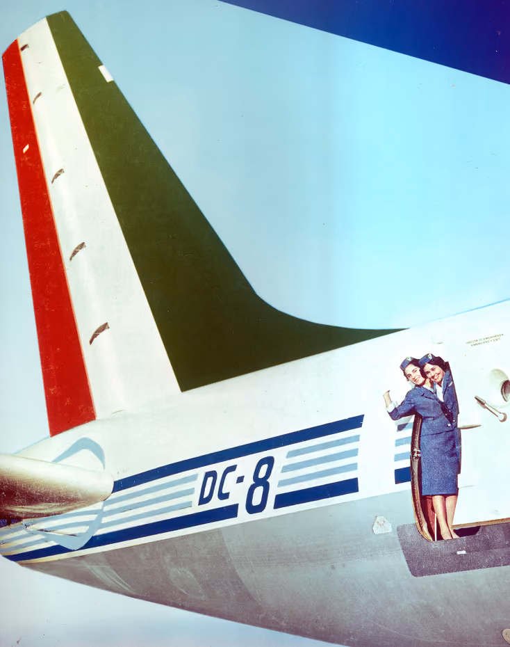 Two flight attendants in vintage uniforms waving from the door of a DC-8 airplane near its tail.