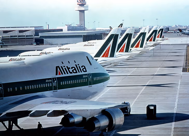 Row of Alitalia airplanes lined up on airport tarmac near terminal and control tower.