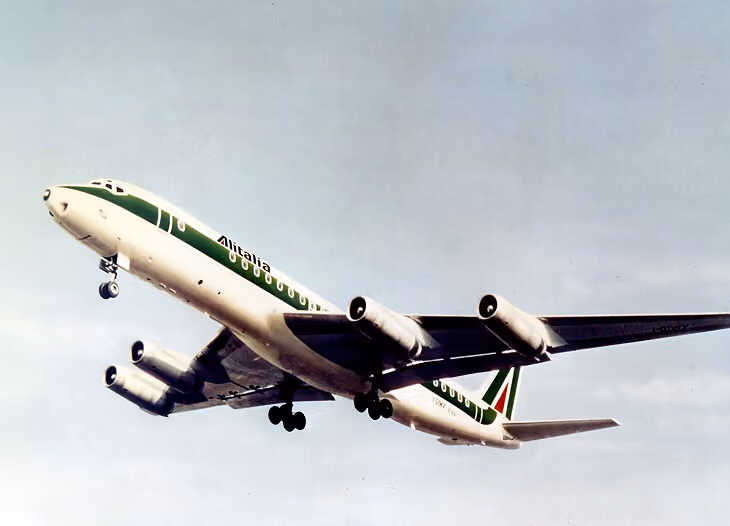 Alitalia commercial airplane in mid-flight with landing gear extended against a cloudy sky.