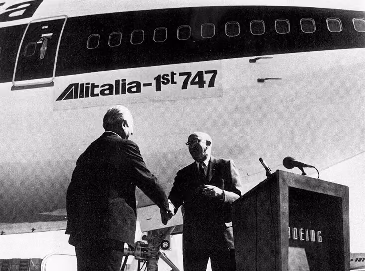 Two men in suits shaking hands in front of an Alitalia 1st 747 airplane with a Boeing podium nearby.